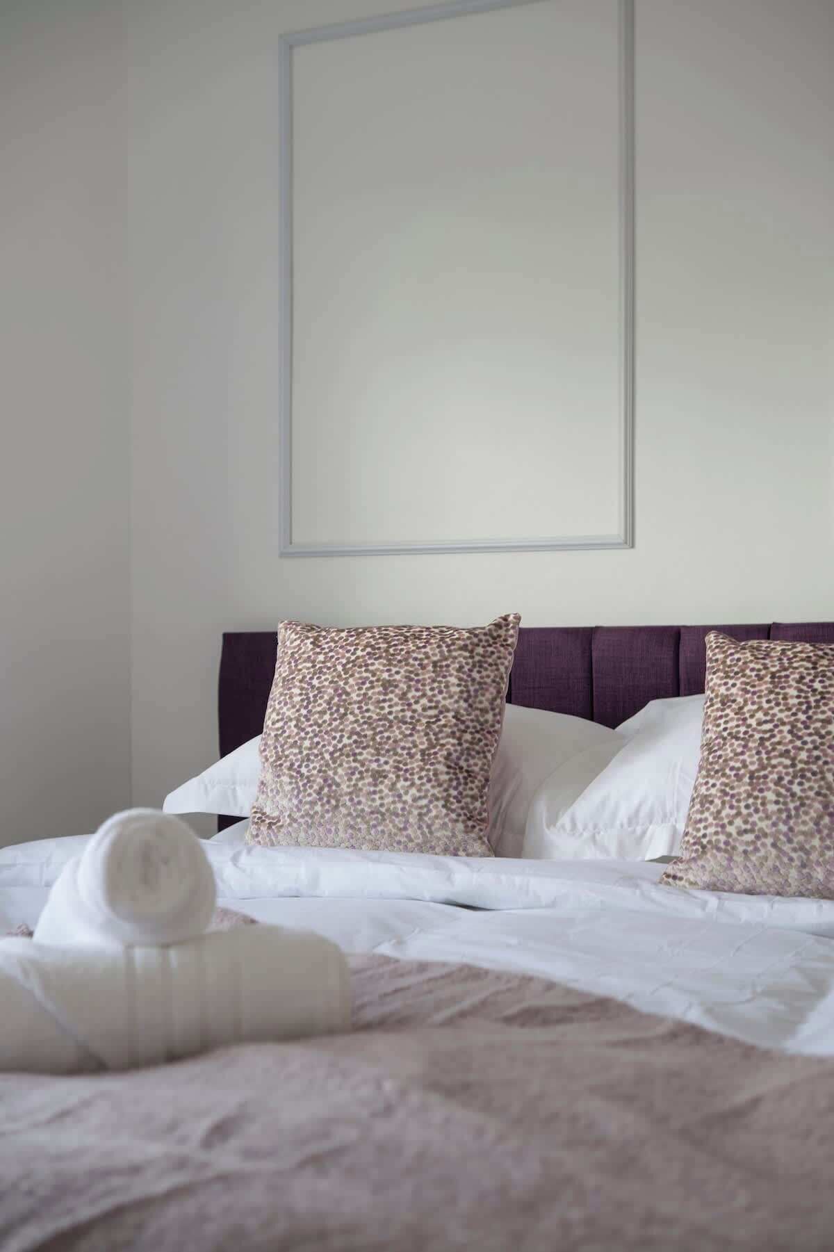 Un lit bien fait, avec du linge de lit blanc et deux coussins à motifs, une tête de lit et un mur sobre.