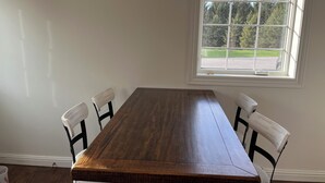 A dining room with a wooden table and white chairs, a window with a view of trees and a road, and a white wall. - Timberside - A Getaway for the Whole Group (Dyersville)