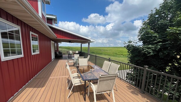 A covered deck with outdoor furniture, a view of a green field, and a red building. - Timberside - A Getaway for the Whole Group (Dyersville)