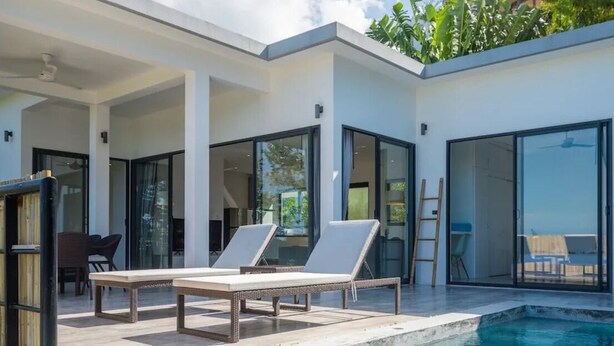 A modern outdoor pool area with lounge chairs and a table, surrounded by glass doors and a white building. - Experience serene sea views, private pool, and tropical luxury in this stunning 3-bedroom villa. (Ko Samui District)