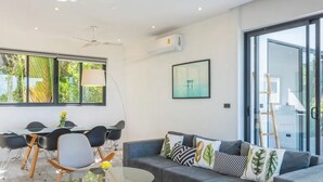 A modern living room with a sofa, dining table, and chairs. There is a large window with a view of greenery outside. - Experience serene sea views, private pool, and tropical luxury in this stunning 3-bedroom villa. (Ko Samui District)
