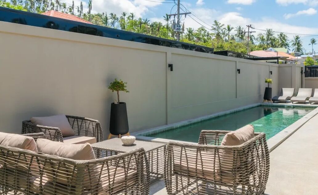 A poolside area with wicker furniture and a small table, surrounded by a low wall and a clear view of the sky and palm trees.