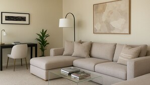 A modern living room with a sectional sofa, a glass coffee table, a desk with a chair, a floor lamp, and a framed abstract painting.