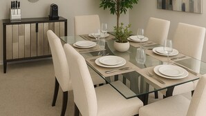 A dining area with a glass table, white chairs, a potted plant, and a black console table with decorative items.