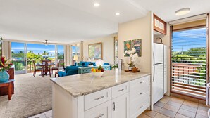A spacious kitchen with a large island, a dining area with a blue sofa, and a view of a lush garden through the sliding glass doors.