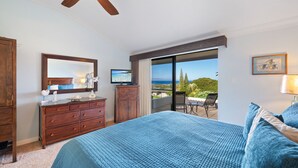 A bedroom with a large bed, a dresser, a television, and a balcony with a view of the ocean.