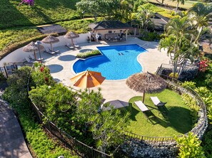 A large swimming pool surrounded by lounge chairs and umbrellas, with a small building and lush greenery nearby. - 2bd chic Island elegance and comfort (Lahaina)