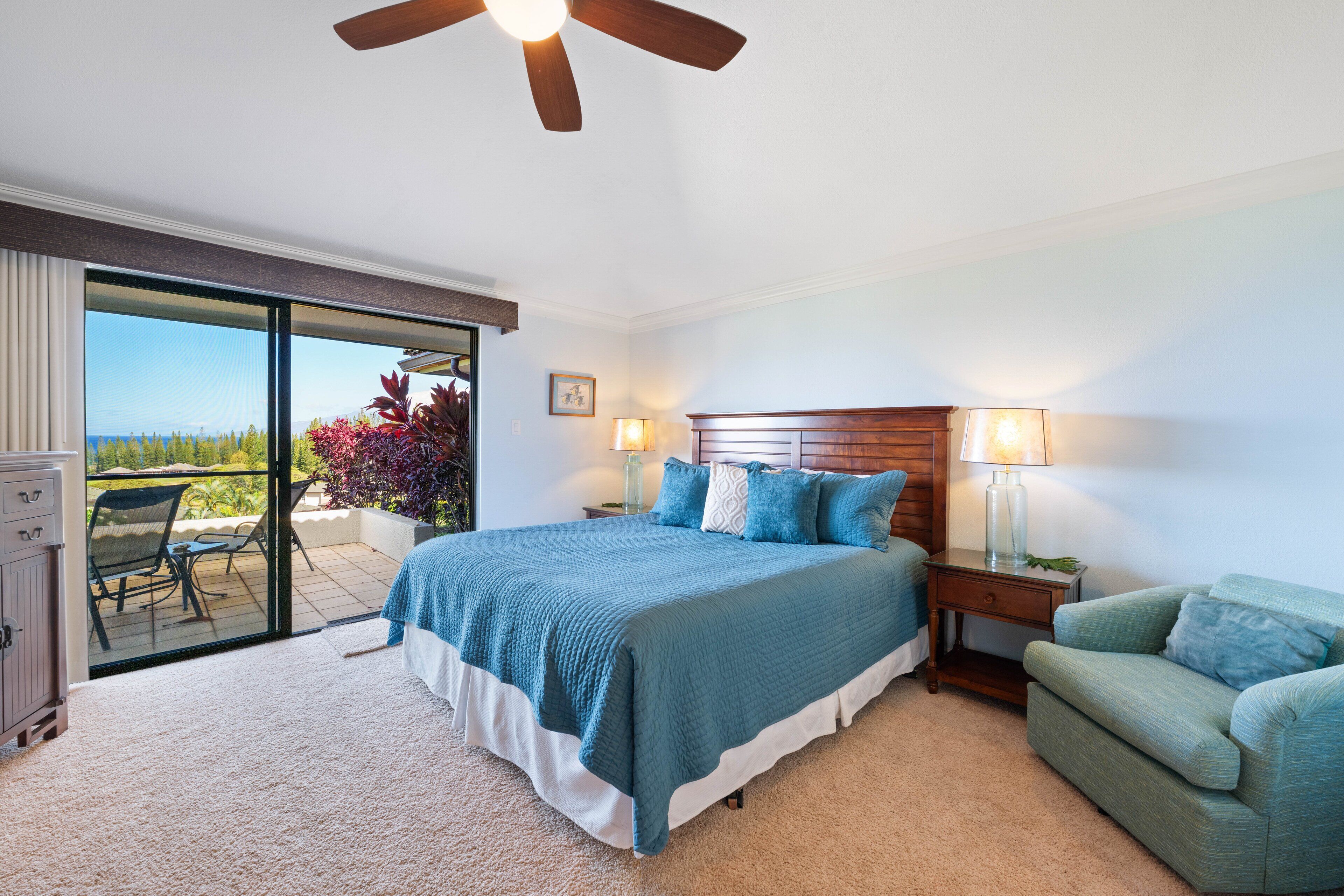 Une chambre à coucher comprenant un lit, une table de chevet, une chaise, un ventilateur de plafond et une vue sur l’extérieur, accessible par une porte-fenêtre coulissante.