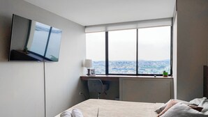 A hotel room with a bed, a desk, a TV mounted on the wall, and a large window offering a city view.