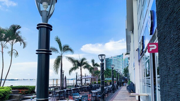An outdoor cafe with tables and chairs, a street lamp, and a view of the water.