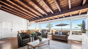 A spacious living area with a wooden ceiling, large windows overlooking a beach, and comfortable seating. - Luxurious Oceanfront Home with Incredible Views (Newport Beach)