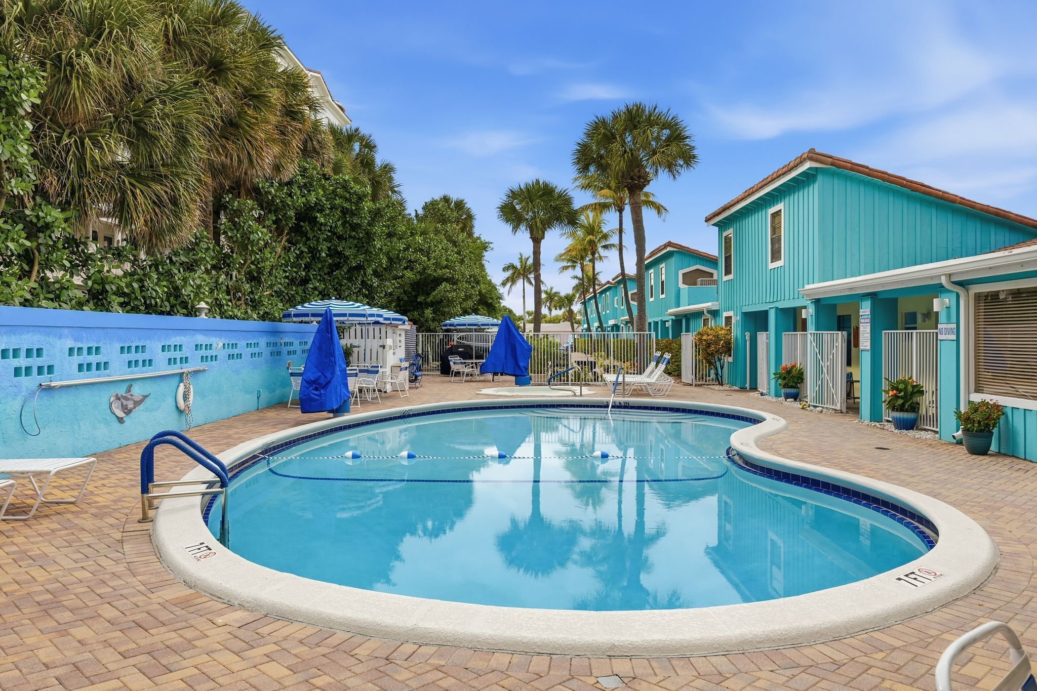 A swimming pool with lounge chairs and umbrellas, surrounded by palm trees and a turquoise building.