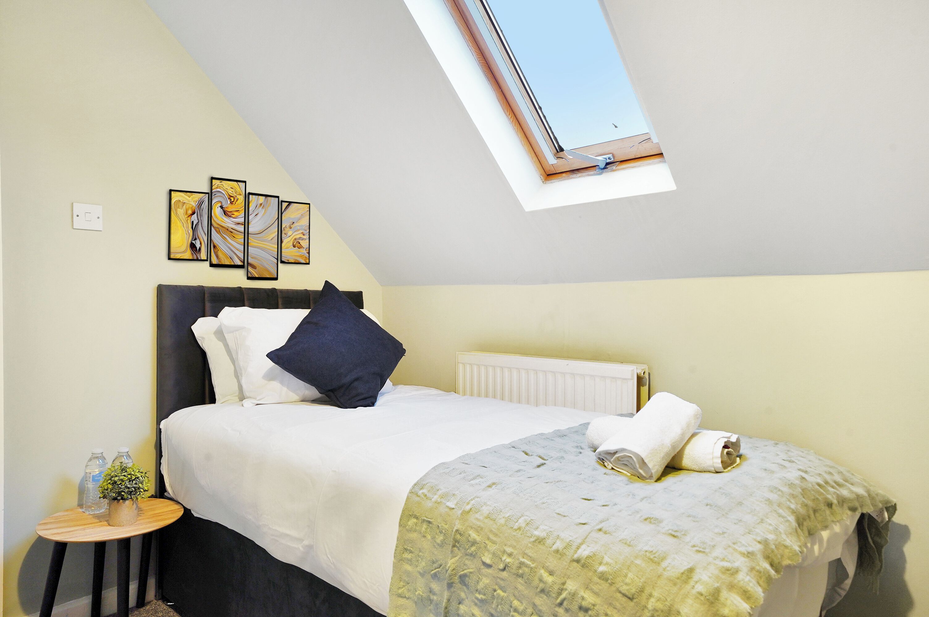 A bedroom with a bed, a small table with a plant, a radiator, and a skylight.