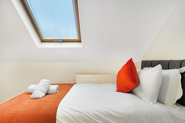 A bedroom with a skylight, a bed with white and orange bedding, and a radiator.