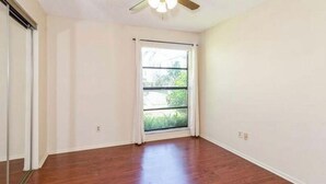 A room with hardwood flooring, a ceiling fan, and a window with white curtains.