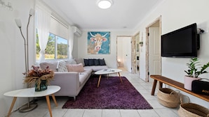 A modern living room with a sofa, coffee table, and a painting of a cow. There is a TV mounted on the wall, a window with curtains, and a purple rug. - Urban Connection Farm (Eagleton)
