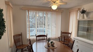 Un comedor con una mesa redonda de madera, seis sillas, un ventilador de techo y una ventana con persianas.