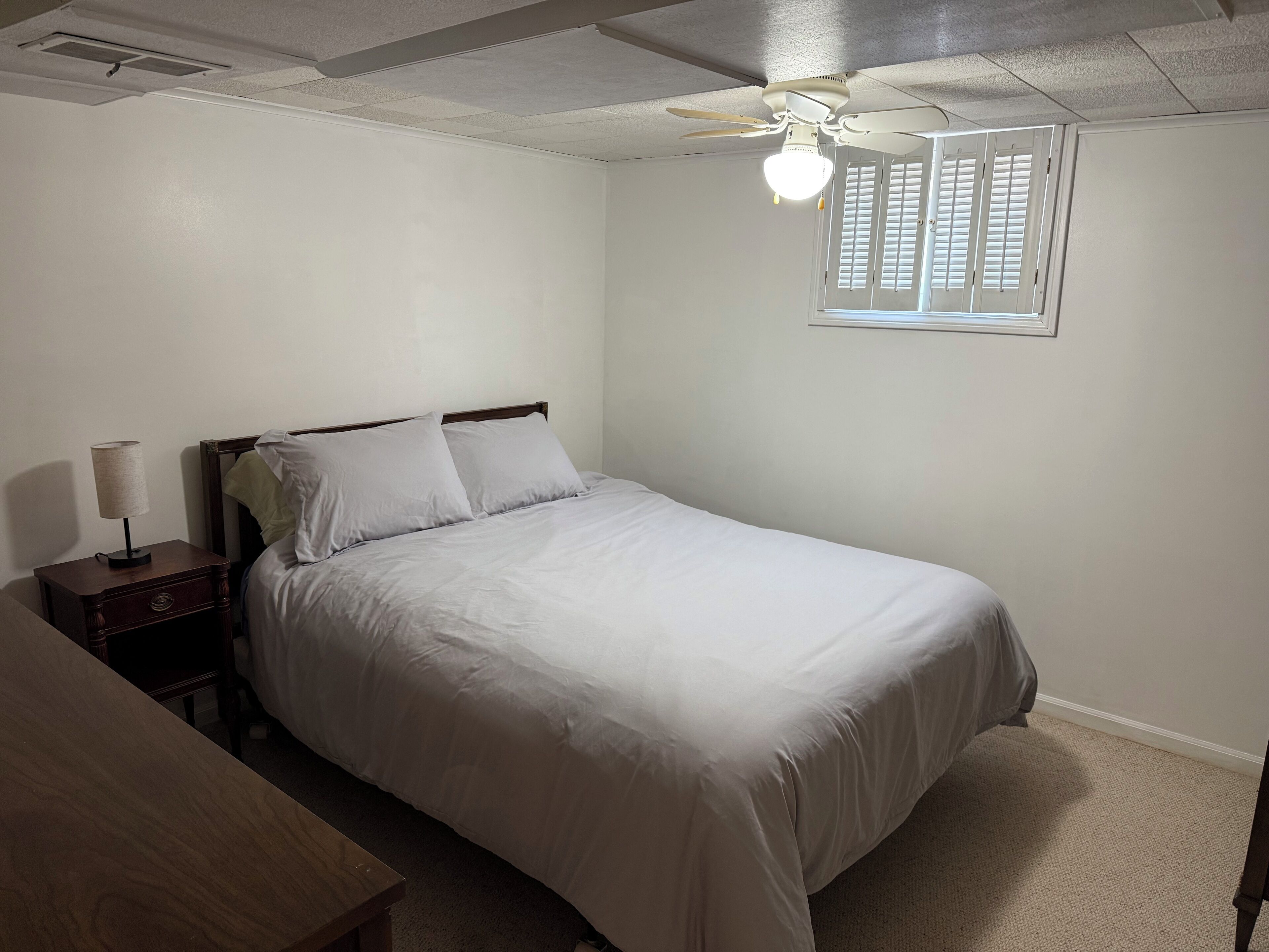 Un dormitorio con una cama, una mesita de noche, una lámpara, un ventilador de techo y una ventana con persianas.