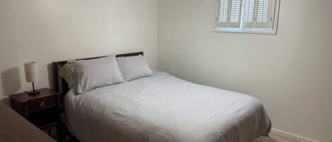 Une chambre à coucher comprenant un lit, une table de chevet, une lampe, un ventilateur de plafond et une fenêtre avec des stores.