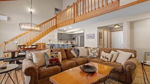 A spacious living room with a large brown sectional sofa, a wooden coffee table, and a wooden staircase leading to an upper level.