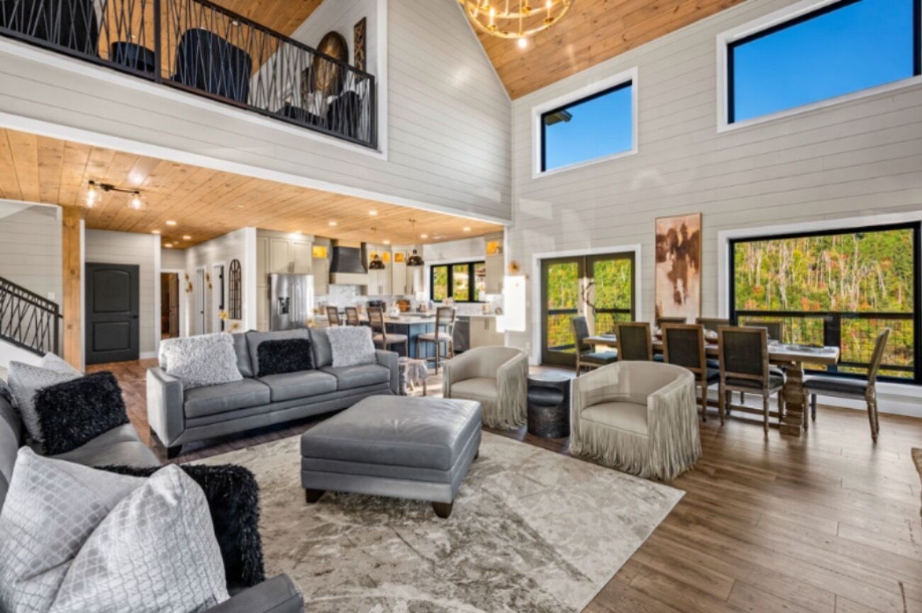 A spacious living area with a grey sectional sofa, a grey ottoman, and a beige armchair. There is a dining area with wooden chairs and a table. The room has large windows with a view of trees.