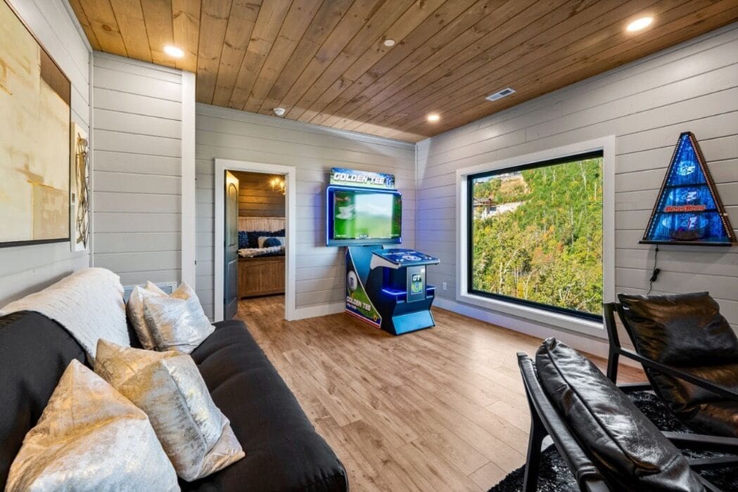 A cozy living room with a black sofa, a gaming console, and a large window offering a view of greenery.