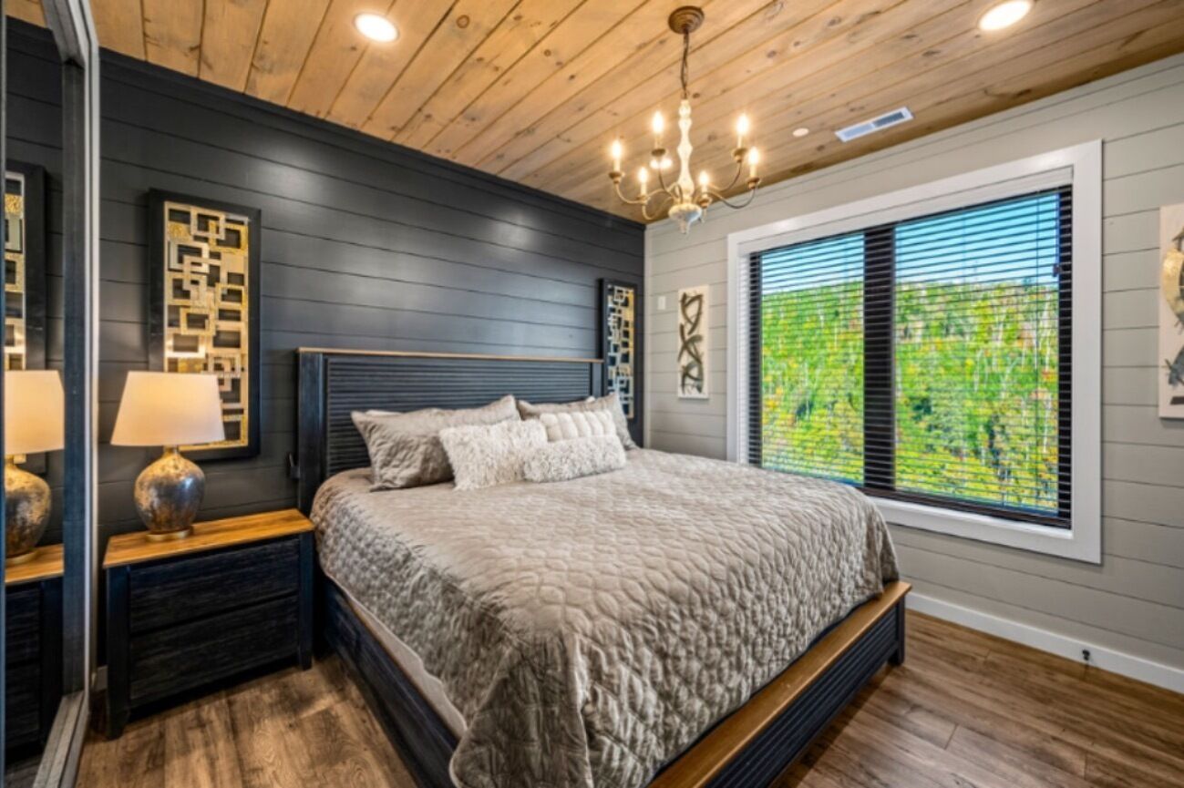 Une chambre avec un grand lit, un plancher en bois, un lustre et une fenêtre avec des stores.