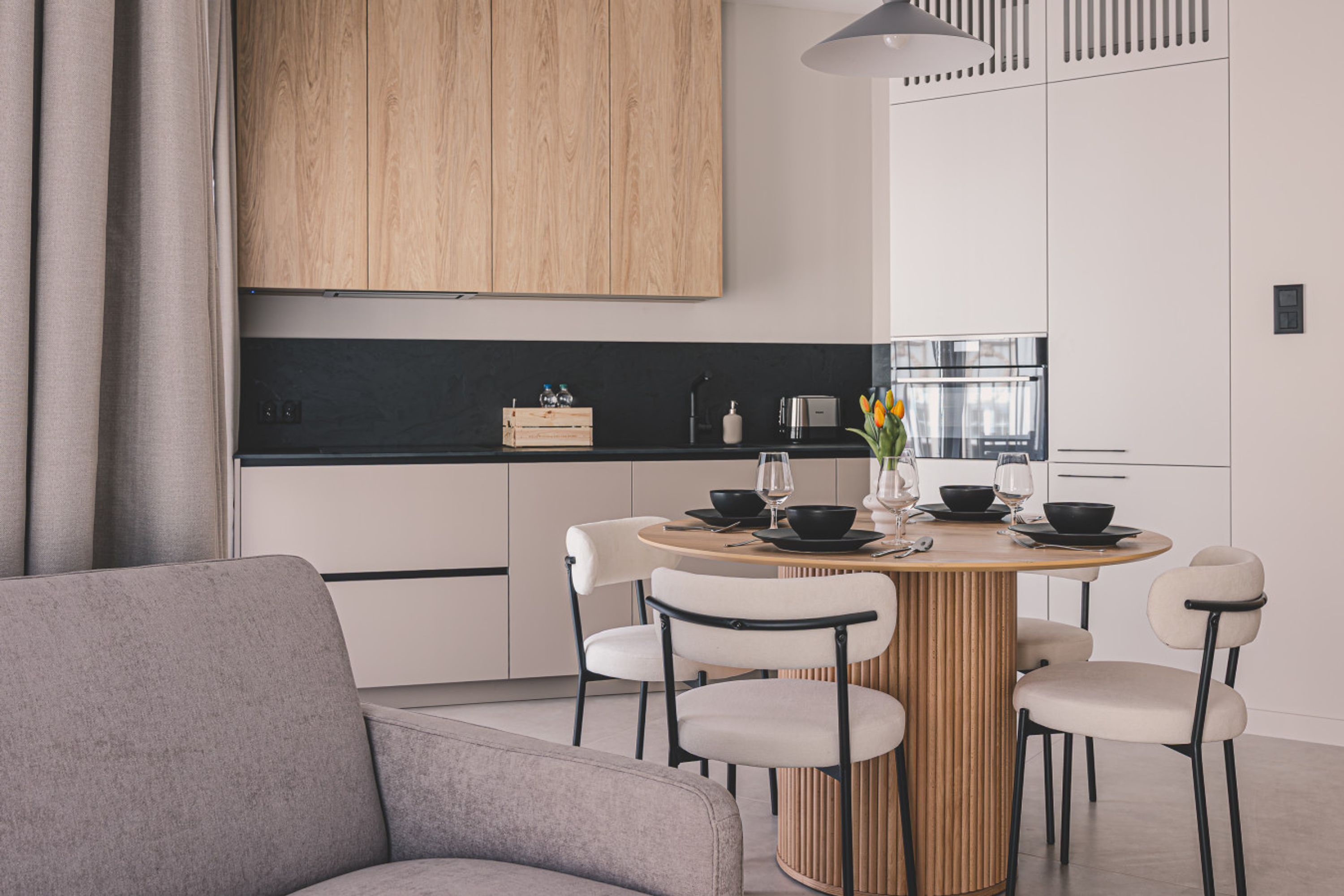 A modern kitchen with a round wooden table, white chairs, and a grey sofa.