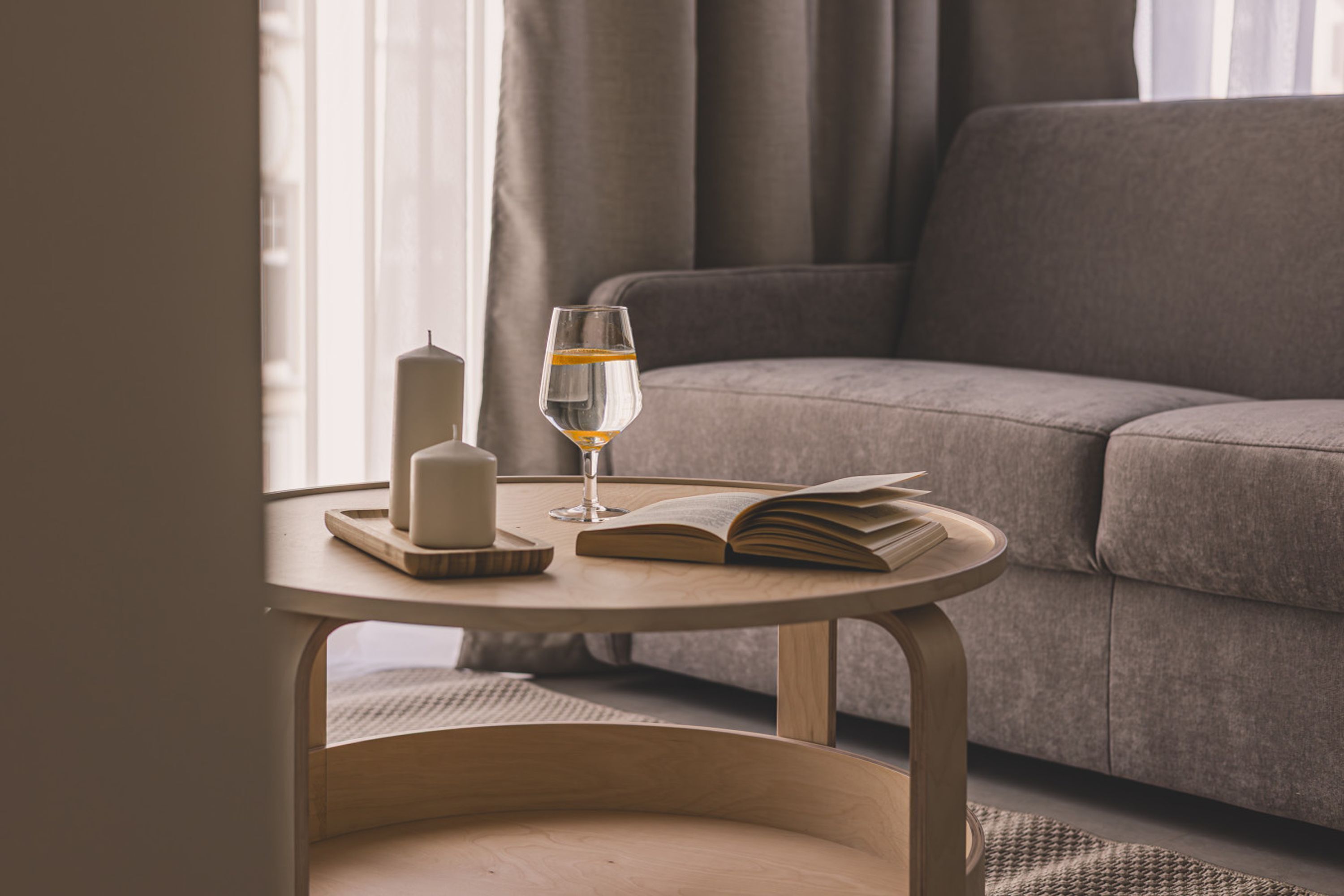 A cozy living room with a round wooden table holding a candle, a glass of water, and an open book.