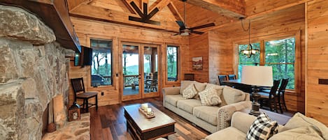 A log cabin interior with a stone fireplace, wooden flooring, a sofa set, and a dining area.