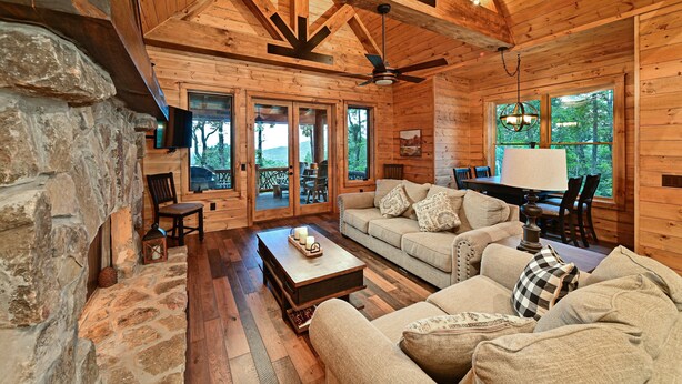 Interior acogedor de una cabaña de troncos con chimenea de piedra, muebles de madera y ventanas amplias que ofrecen vistas al exterior.