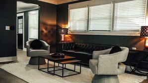 Una sala de estar moderna con una pared oscura, un sofá de cuero negro, un sillón gris, una mesa de centro de madera y dos lámparas de pie.