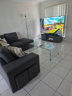 Una sala moderna con un sofá, una mesa de centro de vidrio y un televisor de pantalla plana mostrando un juego de carreras.