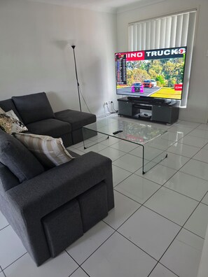 Un salon doté d’un téléviseur à écran plat diffusant un jeu de course, d’un canapé gris, d’une table basse en verre et d’une grande fenêtre du sol au plafond avec des stores.