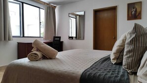 Ein ordentlich bezogenes Bett, ein zusammengerolltes Handtuch, ein Holzschrank, ein Schreibtisch mit Stuhl, ein Spiegel und eine Tür.