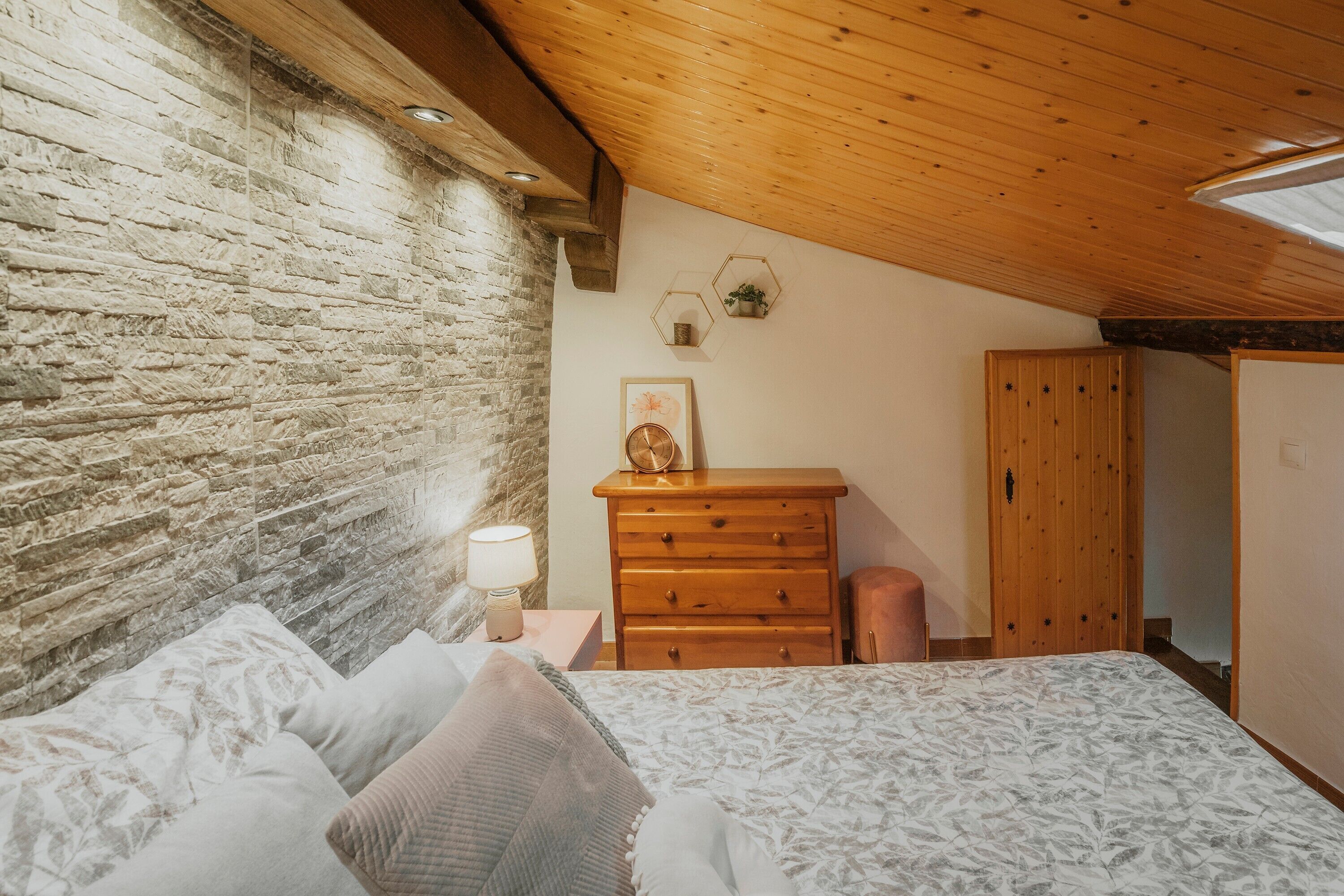 Une chambre avec un mur en pierre, un plafond en bois, un lit avec une literie à motifs, une commode en bois et une table de chevet avec une lampe.