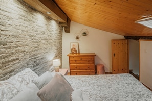 Une chambre avec un mur en pierre, un plafond en bois, un lit avec une literie à motifs, une commode en bois et une table de chevet avec une lampe.