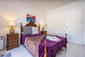 Ein ordentlich bezogenes Bett mit lila Tagesdecke, zwei Nachttischlampen, einem Holz-Kopfteil, einem farbenfrohen Gemälde an der Wand und einer weißen Tür.