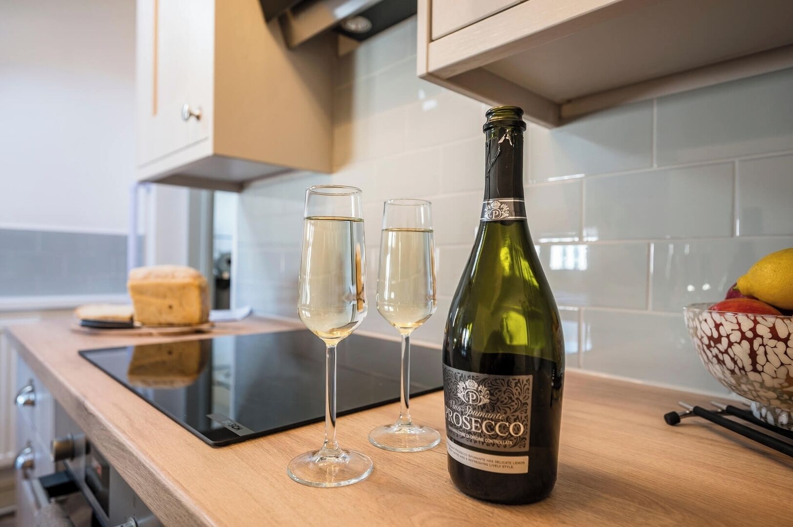 Eine Küchenarbeitsplatte mit einer Prosecco-Flasche, zwei Weingläsern, einem Brotlaib und einer Obstschale.