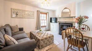 Ein Wohnzimmer mit grauem Sofa, einem gesteppten Ottomanen, einem Esstisch mit Stühlen, einem Kamin und einem Fernseher.