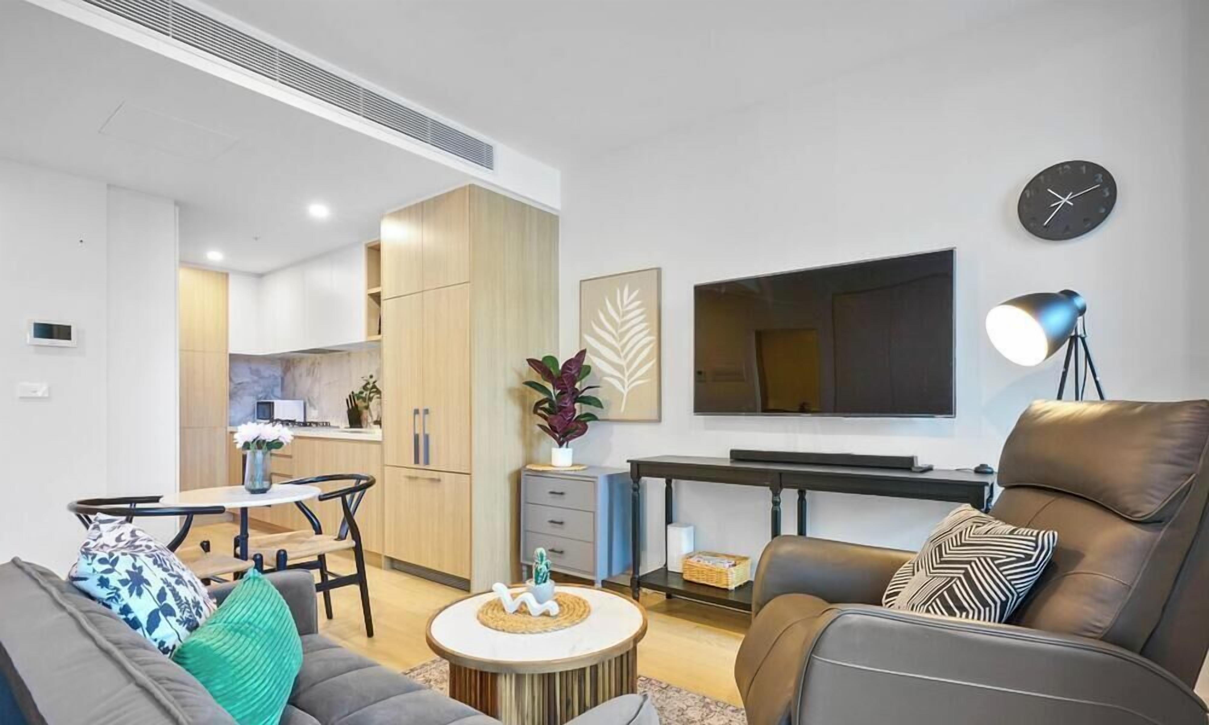 A modern living room with a sofa, coffee table, and a dining area with a table and chairs. A flat-screen TV is mounted on the wall, and there is a lamp on a side table.