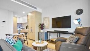 Una sala de estar moderna con un sofá, una mesa de centro y un comedor con una mesa y sillas. Un televisor de pantalla plana está montado en la pared, y hay una lámpara sobre una mesita auxiliar.