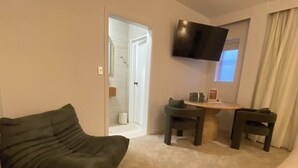 Ein Wohnzimmer mit einer Couch, einem kleinen Tisch, zwei Stühlen, einem an der Wand montierten Fernseher und einer Tür zu einem Badezimmer.