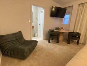 Ein Wohnzimmer mit einer Couch, einem kleinen Tisch, zwei Stühlen, einem an der Wand montierten Fernseher und einer Tür zu einem Badezimmer.