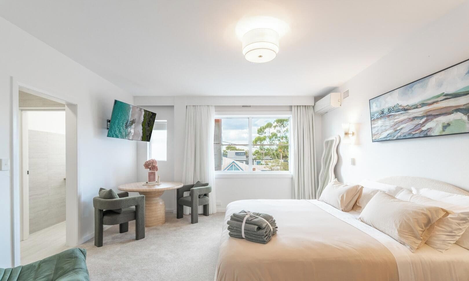 Quarto de hotel com cama, escrivaninha, cadeira, janela com vista e um quadro na parede.