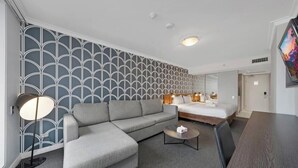 A modern hotel room with a grey sofa, a small coffee table, a bed with a wooden frame, and a large TV on the wall.