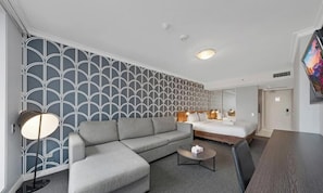 A modern hotel room with a grey sofa, a small coffee table, a bed with a wooden frame, and a large TV on the wall.