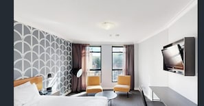 Ein Hotelzimmer mit einem Bett, zwei Sesseln, einem kleinen Tisch, einem Fernseher und einem Fenster mit Vorhängen.