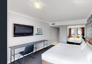 Habitación de hotel con cama, escritorio, silla, televisor y espejo.