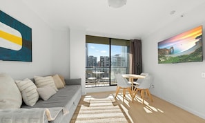 Ein modernes Wohnzimmer mit einer Couch, einem Esstisch mit Stühlen, einem großen Fenster mit Stadtblick und einem Bild an der Wand.